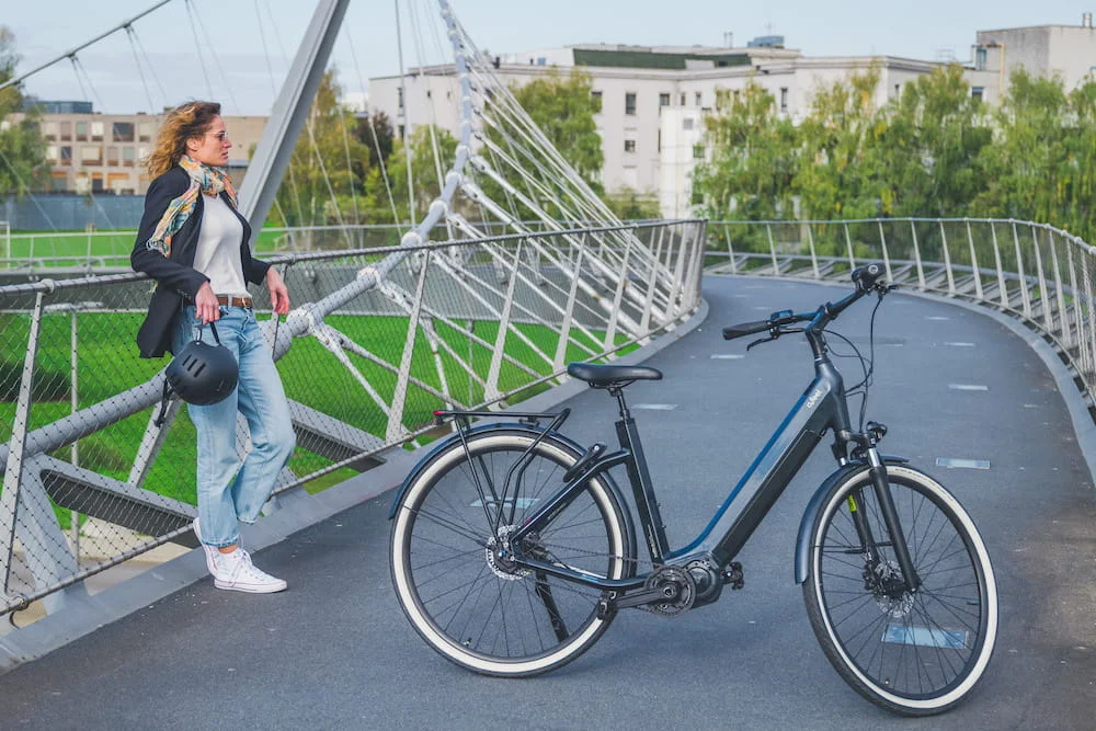 Une femme avec un casque à la main se tient près d'un vélo électrique Urban O2 feel de couleur bleue sur un pont